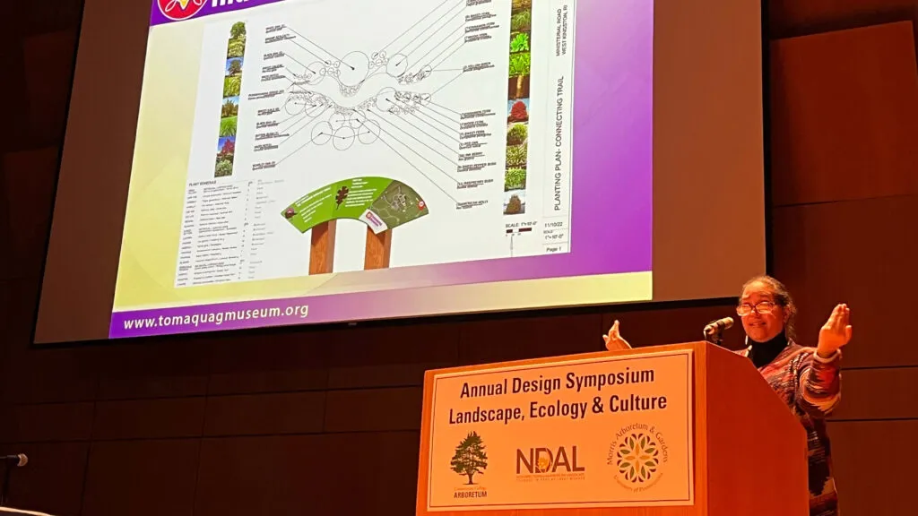 Photo of Lorén Spears from Tomaquag Museum standing behind a podium giving a lecture at NDAL's 2024 Annual Design Symposium. Label on podium reads