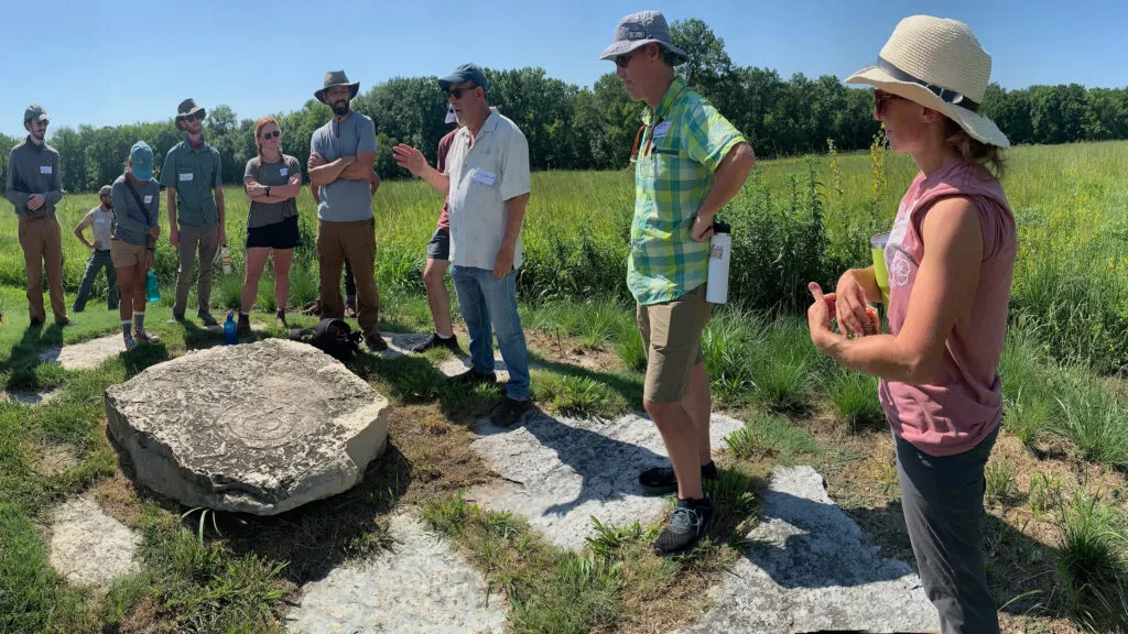 A circle of people standing outside in the sun, listening to Larry Weaner speak. He is standing in the center with his right hand lifted, wearing a baseball hat and sunglasses. There is a meadow behind them and a large, flat rock in front of them.